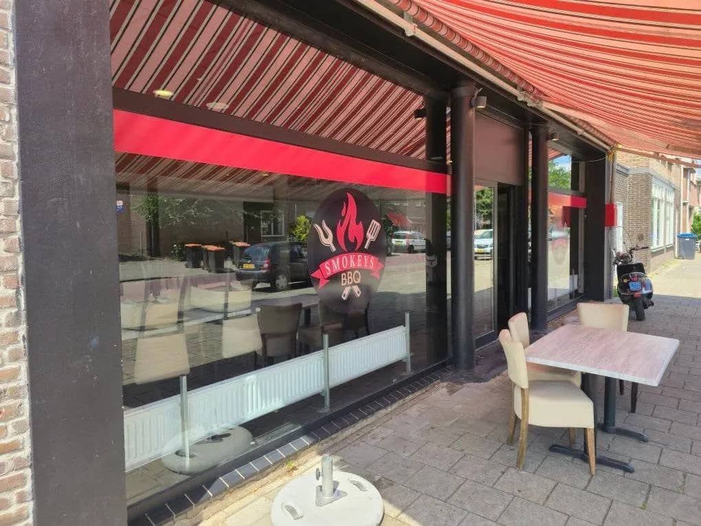 Schoongemaakte glazen pui van een horecazaak met rode zonwering en een terras met tafels